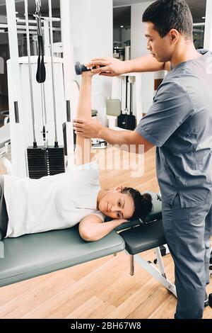 Die Frau in einem Rehabilitationszentrum liegt auf einem Massagetafel und hält eine Hantel in der Hand. Übungen zur Erholung nach einer Verletzung. Rehabilitationstrainer Stockfoto