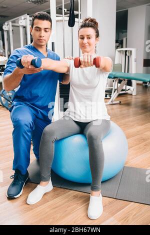 Physiotherapeut, der mit männlichen Patienten in der Klinik arbeitet. Hebt die Hanteln an, stärkt die Armmuskulatur und entwickelt Gelenke Stockfoto