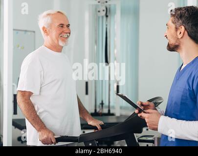 Leitender Patient, der mit seinem Therapeuten im Krankenhaus eine Physiotherapie durchgeführt hat. Ältere Mann Training auf dem Laufband. Stockfoto