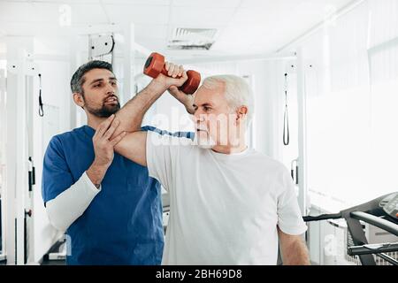 Älterer Mann hebt eine Hantel und macht eine Behandlungsübung mit seinem Physiotherapeuten. Physio-Behandlung im Reha-Zentrum Stockfoto