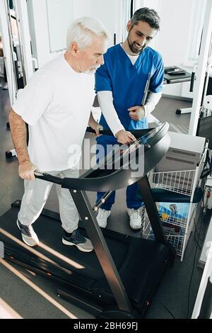 Älterer Mann, der Cardio-Training auf dem Laufband macht, hilft Therapeut älteren Patienten zur Erholung Stockfoto