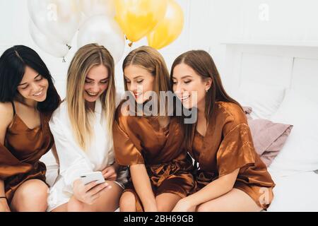 Freundinnen sitzen auf dem Smartphone. Zukünftige Braut mit ihren Freunden feiern eine Junggesellenparty, schauen Sie sich das Foto auf dem Handy Stockfoto