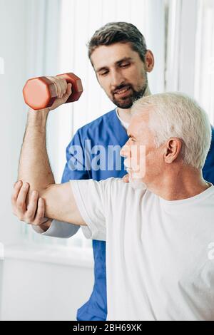 Älterer Mann hebt eine Hantel und macht eine Behandlungsübung mit seinem Physiotherapeuten. Physio-Behandlung im Reha-Zentrum Stockfoto