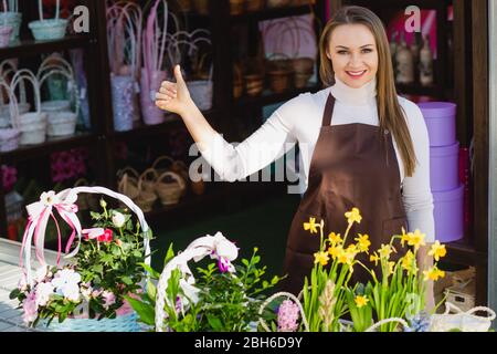 Frau Verkäuferin steht in einem Blumenladen, lächelt und zeigt Daumen hoch. Glückliche Besitzerin eröffnete ihren Blumenladen Stockfoto