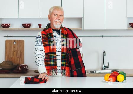 Älterer Mann steht in Oberbekleidung, karierter Schal zu Hause in der Küche, schaut auf die Kamera Stockfoto