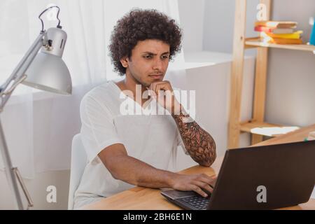 Junger Mann, ein Mulatto mit lockigen Haaren, konzentrierte sich auf Laptop, während er an seinem Arbeitsplatz zu Hause sitzt Stockfoto