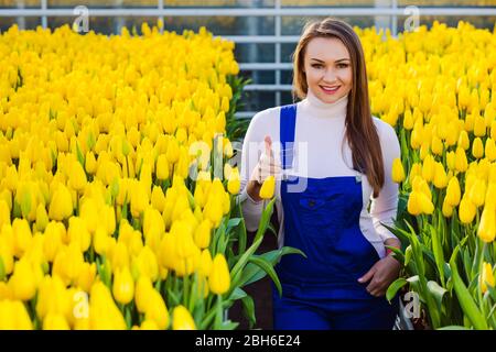 Frau Gärtnerin in Arbeitsuniform gekleidet, lächelnd Blick auf die Kamera, im großen Gewächshaus stehend. Industrielle Kultivierung von Blumen Stockfoto