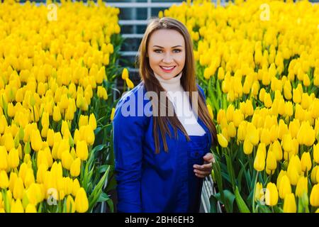 Frau Gärtnerin in Arbeitsuniform gekleidet, lächelnd Blick auf die Kamera, im großen Gewächshaus stehend. Industrielle Kultivierung von Blumen Stockfoto