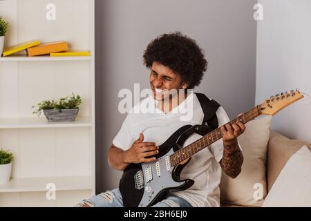 Musiker. Positiv Lockenhaariger Mann, ausdrucksstark spielt die Bassgitarre, zu Hause Stockfoto