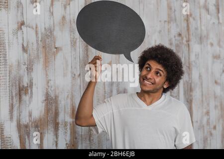 Positive Gedanken. Attraktiver mann Mulatte mit lockigem Haar mit leeren Sprechblasen und es beim Stehen gegen alte Holzwand Stockfoto