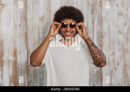 Wählen Sie ein modisches Sommeraccessoire! Portrait Mann mit stilvoller Frisur und perfektem Lächeln Anpassung seiner Sonnenbrille. Im Stehen gegen altes Holz Stockfoto