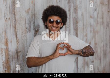 Mit der Liebe. Positive und Curly mulatto Mann, Sonnenbrille tragen, faltete seine Hände in Form eines Herzens, während sich gegen alte hölzerne Wand Stockfoto