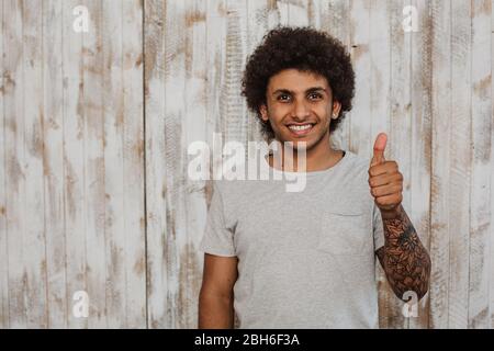 Keine Sorge, seid glücklich! Portrait fröhlich lockig behaart Mann mit perfekten Lächeln zeigt Daumen nach oben. Vor altem Holzhintergrund stehen Stockfoto