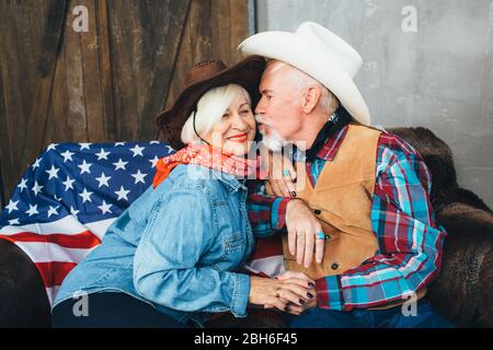 Ältere Paare, in Cowboyhüte gekleidet, lächelnd, sich gegenseitig die Hände nehmend. Hinter, auf der Couch liegt die amerikanische Flagge, die Feier der amerikanischen Stockfoto