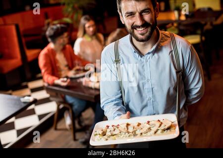 Lächelnder junger Kellner, der Teller mit Fleischgericht im Restaurant trägt Stockfoto