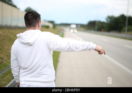 Junger Mann in einem weißen Pullover, der auf der Straße unterwegs ist und versucht, ein Auto anzuhalten. Junge Reisende Anhalter . Anreise mit dem Auto. Stockfoto