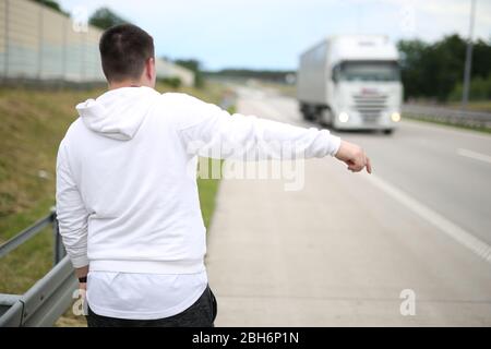 Junger Mann in einem weißen Pullover, der auf der Straße unterwegs ist und versucht, ein Auto anzuhalten. Junge Reisende Anhalter . Anreise mit dem Auto. Stockfoto