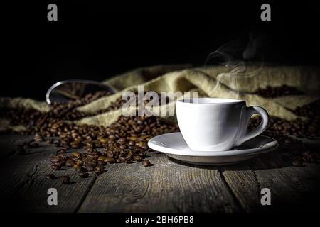 Dampfende Tasse Kaffee auf Holzhintergrund in alten Naturplanken und Sackleinen mit gerösteten Kaffeebohnen. Weiches Licht. Konzept des Getränks mit Kaffee Stockfoto