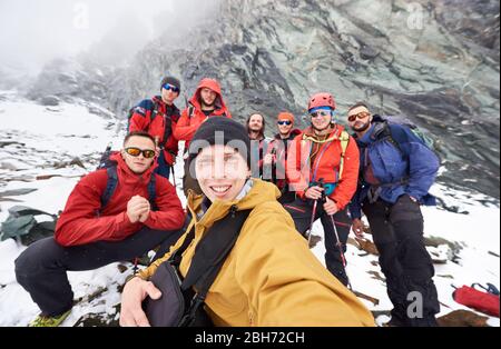 Fröhlicher junger Mann, der mit Freunden Wanderer während der Reise in den Winterbergen fotografiert. Eine Gruppe männlicher Reisender, die die Kamera angeschaut und lächelt. Konzept des Reisens, Wanderns und Fotografierens. Stockfoto