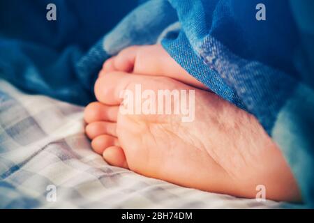 Die Beine eines schlafenden Mannes liegen auf einem karierten Laken auf dem Bett, bedeckt mit einer leuchtend blauen Decke und am Morgen durch Licht beleuchtet. Stockfoto