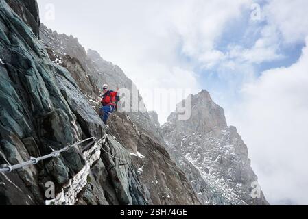 Seitenansicht des männlichen Alpinisten in Sonnenbrille und Schutzhelm, der Fixseil hält, während er den Berg aufsteigt. Bergsteiger zeigt Siegerbewegung und schaut mit einem Lächeln auf die Kamera. Konzept des Bergsteigens Stockfoto