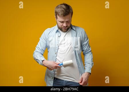 Ruhiger, fokussierter Mann reinigt seine Kleidung von Tierhaaren mit einer Rolle mit Klebeband, auf einem gelben Hintergrund. Stockfoto