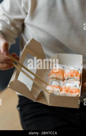 Mann essen Rolle philadelphia mit Garnelen Essstäbchen aus Handwerk-Box. Lieferservice japanische Lebensmittel Rollen in Handwerk-Box Stockfoto