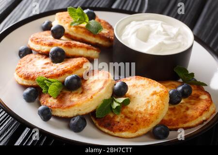 Ukrainisches Frühstück von Hüttenkäse Pfannkuchen mit Heidelbeeren, Minze und saure Sahne Nahaufnahme in einem Teller auf dem Tisch. Horizontal Stockfoto