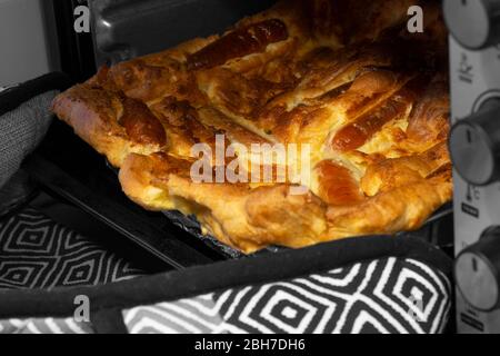 Kröte im Loch, Yorkshire Pudding mit Schweinewurst, die von einer Person mit Ofenhandschuhen aus dem Ofen genommen wird. Selektives Farbbild Stockfoto