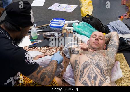 Ein Mann, der auf der 12. London Tattoo Convention 2016, Tobacco Dock, 50 Porte, ein Arm-Tattoo von einem Tätowierer mit der Rake & Stamping Stick Technik bekommt Stockfoto