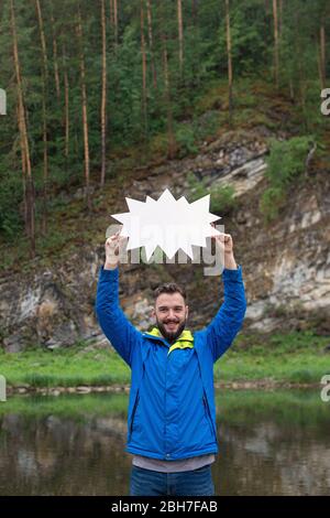 Junger lächelnder Mann, der am Ufer des Flusses steht und ein weißes Brett in den Händen über seinem Kopf hält, felsige Klippen auf dem Hintergrund. Sommer ist die beste Zeit Stockfoto