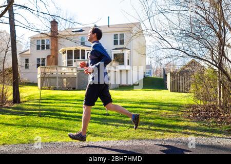 Junger Mann Joggen in Bewegung auf dem Weg in Wohnviertel im Frühjahr mit Haus Hinterhof im Hintergrund Stockfoto