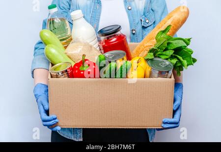 Frau in Handschuhen hält Spendenbox Lebensmittel für Menschen in Isolation auf gelbem Hintergrund. Wesentliche Güter: Öl, Konserven, Getreide, Milch, Veg Stockfoto