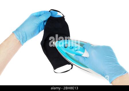 Männliche Hände in blauen Einweg-Nitril-Handschuhe bügeln eine schwarze wiederverwendbare Gesichtsmaske. Waschbare Stoffmaske Hygiene. Erste Person Ansicht, isoliert auf weiß. Stockfoto