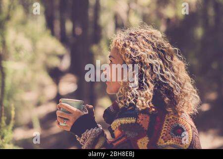 Schöne Frau im Herbstwald das Gefühl mit der Natur mit einem warmen Pullover genießen - Dame sitzt mit Bäumen im Nackgrund in einem Wald und hält eine Tasse mit einem heißen Getränk in den Händen. Mädchen Reise und Fernweh Millennial Konzept Stockfoto