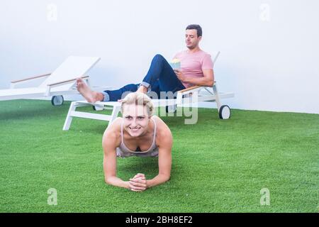 Fröhlich fit Frau tut Plank zu Hause im Garten auf dem grünen Teppich - Freund im Hintergrund Junk Food essen und ruhen auf dem Stuhl - tausendjährige verschiedene Lebensweise für nettes Paar Stockfoto