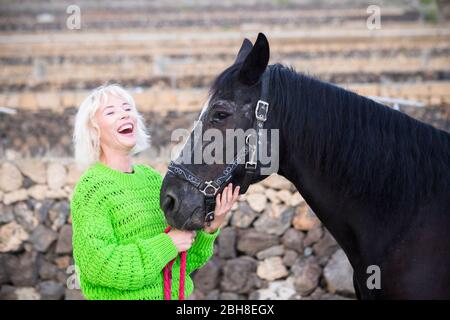 Blonde Mädchen lachen viel mit ihrem besten Freund schwarz schönes Pferd. Farben und Happy Lifestyle Konzept mit Haustieren Therapie in Outdoor-Freizeit-Aktivität - fröhliches Konzept Stockfoto