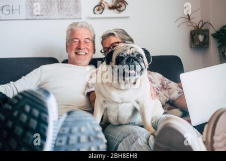 Kaukasische ältere Paar sitzen auf der Couch mit lustigen Mops Hund suchen. Glück mit Liebe zu Tieren und beste Freund Konzept - Home Alltag mit netten Menschen und alten Welpen