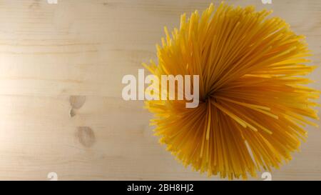 Top-down-Wohnung Blick auf eine Handvoll Spaghetti gespenkte Flecken, die wie eine gelbe Blume auf einem hellen Holztisch Hintergrund italienischen Küche c aussehen Stockfoto