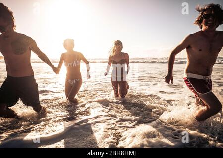 Gruppe von schönen Millennial Alternative junge Menschen Spaß haben und genießen Sie den Sommerurlaub zusammen in Freundschaft im Wasser des Ozeans mit Sonne im Hintergrund laufen Stockfoto