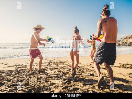 Gruppe von Menschen junge kaukasische Männer und Frauen spielen mit Wasserpistole am Strand während der Freunde Urlaub zusammen im Freien in einem sonnigen Urlaubstag - aktive Jugendliche Alternative Millennial Stockfoto