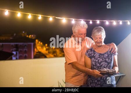 Happy Senior Erwachsene Paar mit einem Schokoladenkuchen nach dem Abendessen zu Hause im Freien auf der Terrasse mit Party Glühbirne Licht - Geburtstagskonzept oder Neujahr Feier für Erwachsene Menschen - Stockfoto