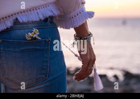 Gruppe von Gänseblümchen in der Gesäßtasche von weiblichen Jeans von hinten gesehen - genießen Sie Leben und Lifestyle-Konzept - Ozean und Sonnenuntergang im Hintergrund - Nahaufnahme Mode Hippie-Stil Stockfoto