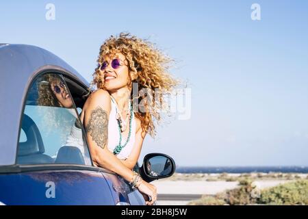 Schöne fröhliche fröhliche kaukasische Frau außerhalb des Autos mit Wind in lockig langen blonden Haaren - attraktive Menschen genießen die Freiheit im Freien Küste Meer Urlaub Ort Stockfoto