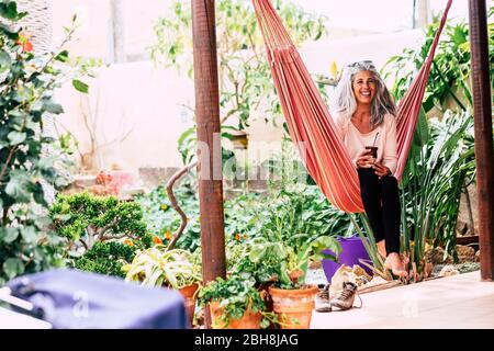 Fröhliche lächelnde Vielfalt Menschen Konzept mit schönen trendigen erwachsenen Frau mit weißen langen Haaren lachend setzen Sie sich auf eine Hängematte zu Hause im Garten trinken einen Tee Stockfoto