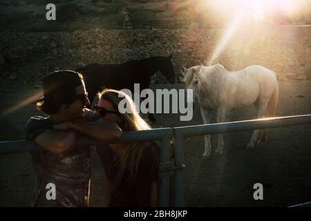 Romantisches Licht und Paar, das mit Liebe beim Sonnenuntergang mit zwei schönen Pferden im Hintergrund flüstert - Romantik und Beziehungskonzept im Freien mit Sonnenlicht Stockfoto