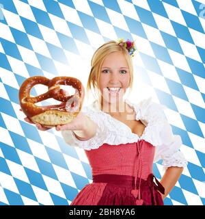 Lachende junge Frau in Dirndl hält eine Brezel vor der bayerischen Flagge Stockfoto