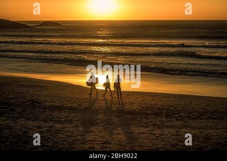 Bei Sonnenuntergang wandern drei Personen mit einem Bodyboard am Strand Llandudno entlang, in der Ferne sind Surfer Stockfoto