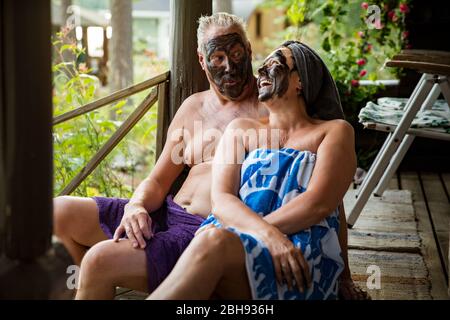 Gealtertes Paar mit schwarzer Gesichtsmaske nach finnischer Sauna. Reifer Mann und Frau in Handtüchern sitzen auf Holztreppen, entspannen, lachen und Spaß haben. Stockfoto