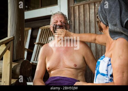 Gealtertes Paar mit schwarzer Gesichtsmaske nach finnischer Sauna. Reifer Mann und Frau in Handtüchern sitzen auf Holztreppen, entspannen, lachen und Spaß haben. Stockfoto